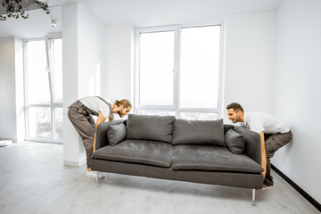 Two professional movers in workwear placing large couch in the living room of the modern white apartment. Furniture delivery concept