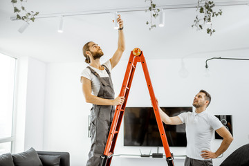 Two repairmen or professional electricians installing light spots, standing on the ladder in the...