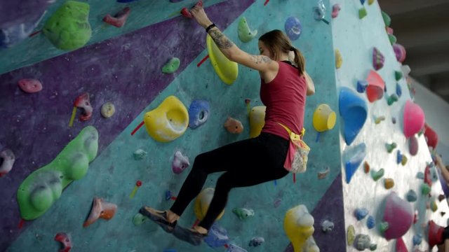 Tracking shot of skilled Caucasian female climber bouldering on artificial wall at indoor climbing gym, swiftly moving around various foot and hand holds and hanging off slope