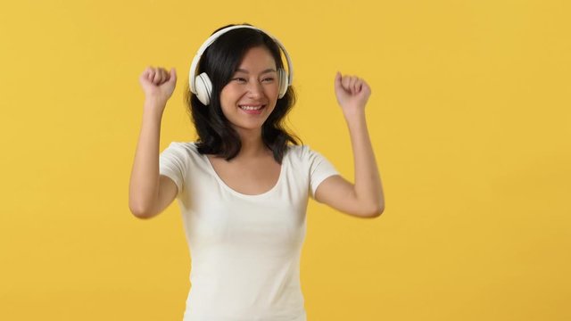 Young happy smiling Asian woman in white t-shirt listening to music on headphones and dancing against yellow background