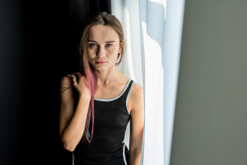 young pretty emotional and sexy girl with blue eyes in a black dress sits by the window, a sunbeam...