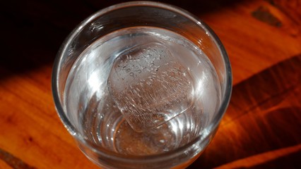 Top view photo of a glass full of clear water, with an ice cube in it, on a wooden table.