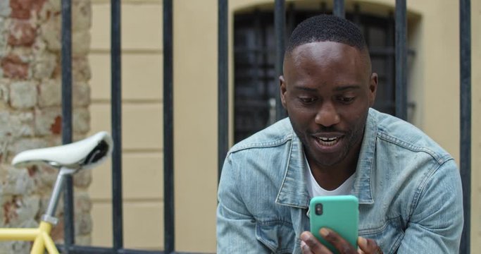 Close Up of Smiling African American Man Having a Video Call via his Smartphone. Male Tourist Waving, Smiling while Sitting near Stylish Bike with Old Metal Fance at the Background. Front View.
