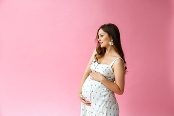 Studio shot of happy pregnant woman dressed in comfort home dress on a pink background, touching gently her belly and smiling at camera. Pregnancy healthy motherhood concept.