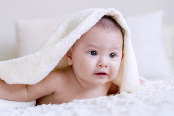 Cheerful cute baby looking at camera under white blanket looking at something. Innocence baby crawling on white bed with towel on his head at home.