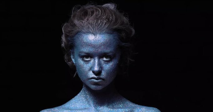 girl with a blue face in sparkles close-up against a black wall looks at the camera