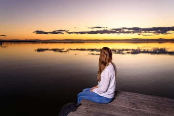 Fototapeta na wymiar Long-haired young woman relaxing admiring the golden sunset on a beautiful pier by a natural lake.