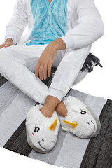Cropped medium shot of a man in white velour pyjamas and plush house slippers made in the form of white smiling unicorn. The man is sitting on the striped carpet and a gray plaid.