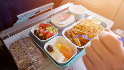 Lunch in plane: bread, meat with vegetables and rice, pieces of cheese, dried apricot, salad and beverage in cup. Woman is eating in airplane during flight food served on tray, hand with fork closeup.