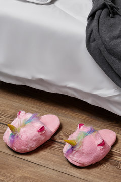Subject shot of plush house slippers made in the form of pink unicorn with rainbow mane. The slippers are next to the bed with white linen, pillow and a gray plaid on it. 
