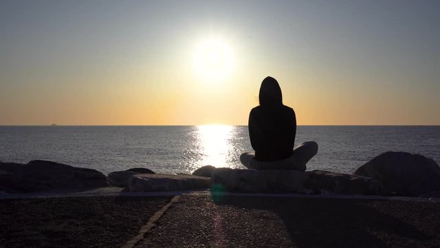 Silhouette of a girl in hoody enjoying early morning sunrise off Puerto Banus pier overlooking the Mediterranean sea, yoga pose Bound Angle Pose (Baddha Konasana), shot in 4k on gimbal