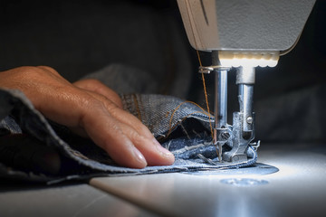 Hand of the seamstress is using a white industrial sewing machine to sew the seams of blue jeans.