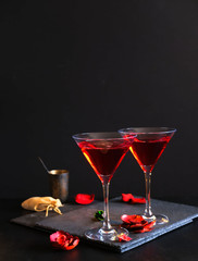 Two cosmopolitan cocktails in a glass. Dark luxury background. In the background an old silver bowl, a bag of tobacco and scattered rose petals.