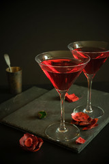 Two cosmopolitan cocktails in a triangular glass. In the background an old silver bowl and scattered rose petals. Dark luxury background.High resolution.