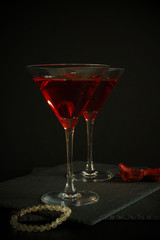 Two cosmopolitan cocktails in a triangular glass. Nearby is an out of precious stones bracelet and scattered rose petals. Dark luxury background.