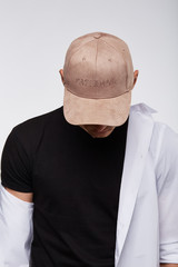 Cropped upward shot of a dark-haired man, wearing beige velvet baseball cap with lettering...
