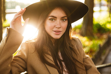 Portrait of stylish young brunette woman sitting on bench in autumn park