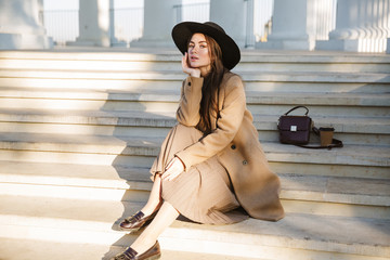 Portrait of adorable young brunette woman sitting on building stairs outdoors