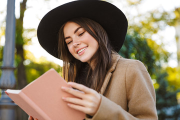 Portrait of joyful woman wearing coat and hat reading book outdoors