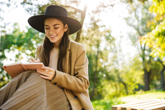 Image of joyful woman holding diary book while sitting on bench in park