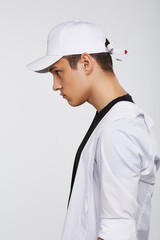 Cropped side photo of a dark-haired man, wearing white baseball cap with embroidery red heart on the strap on the back side of a cap, black t-shirt and white shirt. He is posing on grey background