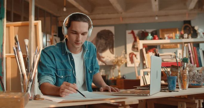 Young Caucasian handsome guy in headphones painter drawing a sketch with a pencil and depicting a picture from the screen of the laptop computer.