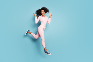 Fototapeta na wymiar Full length photo of amazing dark skin curly lady jumping high walking down street showing v-sign symbols wear pink shirt trousers isolated blue color background