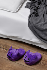 Subject shot of purple plush house slippers made in the form of the smiling devil emoji. The slippers are next to the bed with white linen, pillow and a gray plaid on it. 