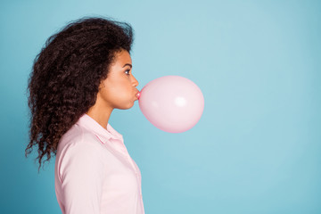 Closeup profile photo of pretty dark skin lady holding big air balloon in mouth making decorations for surprise party wear pink shirt isolated blue color background