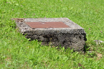 Heavy rusted metal manhole cover on top of old concrete foundation in local field surrounded uncut green grass on warm sunny summer day