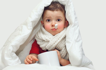 A little cute boy gets sick and measures the temperature with an oral thermometer and sits covered with a blanket and with a large knitted scarf around his neck.