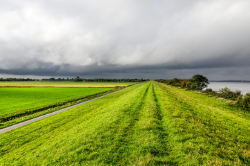 Fototapeta na wymiar Dramatic dark clouds over a vast landscape on the island of Schouwen-Duiveland, The Netherlands, with a grassy dike, green fields and lake Grevelingen