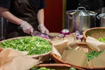 Cook's hands in cellophane gloves cutting red onion into thin slices. Cooking vegetable ingredients. Against the background of lettuce leaves and pans made of stainless steel.