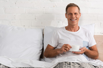 Man holding coffee cup in bed and smiling at camera