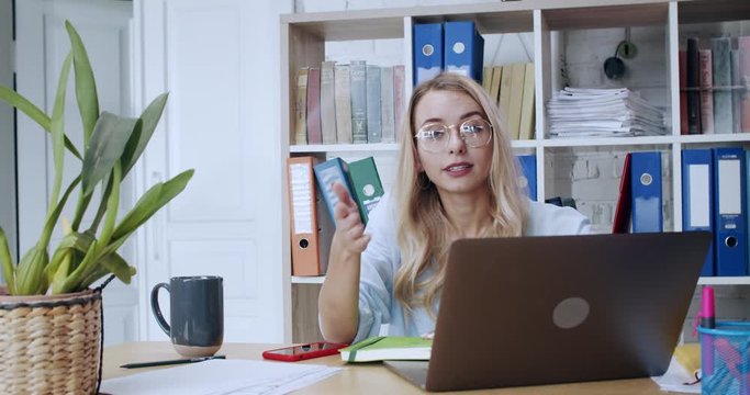 Blonde young businesswoman sitting by laptop in the office talking at camera explaining work or services. Portrait of ambitious talkative female employee.