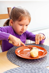 Primary school young girl sitting on a chair near a table, eating potato dumplings all by herself, holding a fork in her hands cutting the food. Child using cutlery concept, finishing her meal