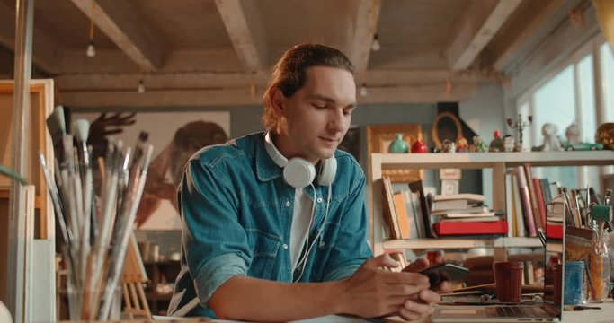 Close up of the cheerful Caucasian young guy painter sitting at the table in his working workshop and chatting on his smartphone while tapping and texting.