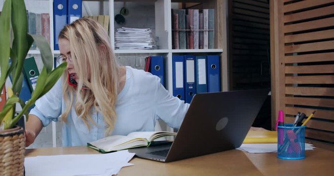 Female office secretary in a hurry is calling on smartphone searching things on desk. Irritated woman employee using laptop doing multi-tasking at office workspace.