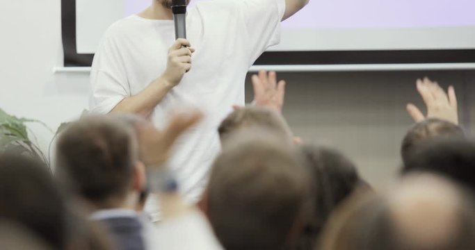 Unrecognizable man speaker talking in large audience rising hand and viewers repet after him, view from audience. Public speech concept. Business educational conference, seminar, training in group.