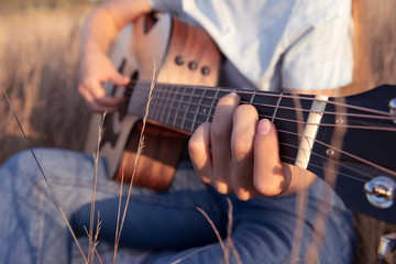Girl plays the acoustic guitar in the field on sunset. Fingers on guitar fretboard. Nature and music. Closeup. - obrazy, fototapety, plakaty