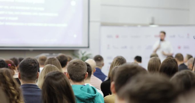 Viewers in large audience photographing man speaker on smartphones at business eductional training, back view. Unrecognizable man talking microphone showing presentation on screen, blur background.