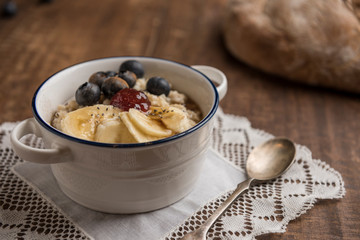 Porridge with banana, oat flakes, blueberries, chia, cinnamon, maple syrup and strawberry jam on rustic wooden table. Country loaf in the background. Horizontal. Selective focus