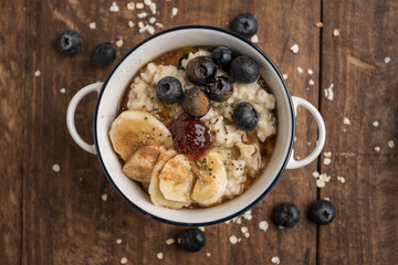 Top view of vegan porridge on wooden table with oat flakes, banana, blueberries, chia, cinnamon, maple syrup and strawberry jam. Horizontal.