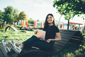 Portrait of cheerful female freelancer taking rest at wooden modern bench in public city park with...