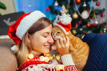 Christmas celebration with cat. Woman playing and hugging pet in Santa's hat by New year tree at home.