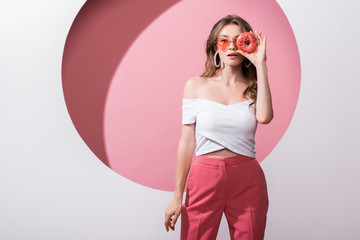 attractive girl covering face with tasty doughnut and standing on pink and white