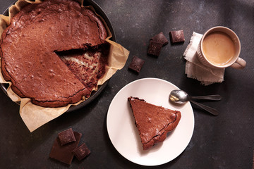 Dark chocolate dessert - homemade brownie cake with pieces of dark chocolate, cup of coffee, spoons on dark rustic background. Top view, flat lay delicious breakfast. 