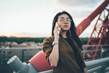 Serious hipster girl in spectacles making international cell conversation for discussing roaming information during talking with international operator, Asian woman pondering on received news