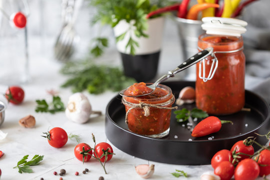 A sauce of fresh red tomatoes in a glass jar on a black plate. Sprig of fresh cherry tomatoes, garlic, hot pepper, dill and parsley on a white table. A can of homemade ketchup.Close up