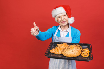 Portrait of pretty charming cheerful aged woman with wrinkle showing gesturing sweet homemade bakery products isolated on red background, christmas recipes.